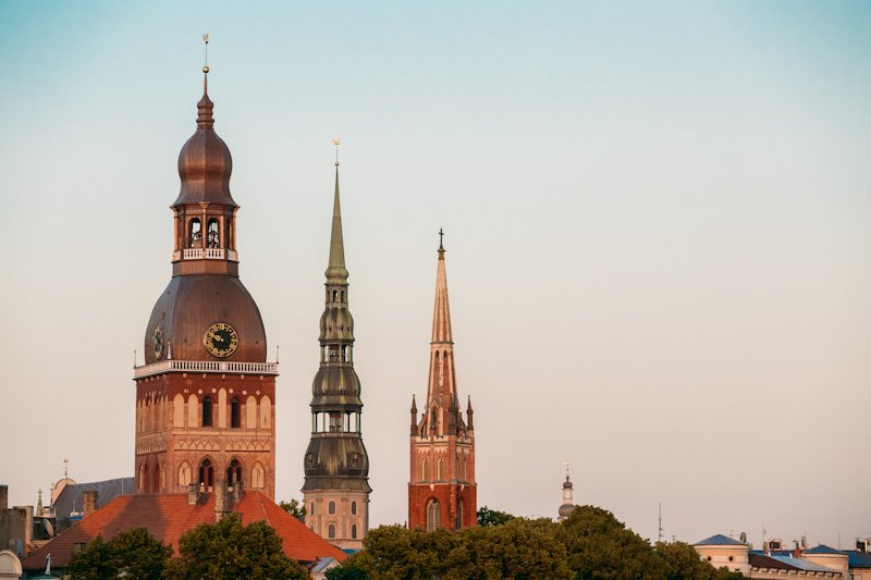 Riga, Latvia. Close View Of Three Towers With Steeples Of Riga Cathedral, St. Peter's Church And St. Saviour's Anglican Church In Old Town In Summer Under Blue Sky.